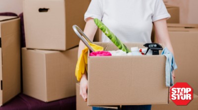 Alt text: Person carrying a box of cleaning supplies and household items while spring cleaning and organizing for self-storage, surrounded by moving boxes in a NYC apartment.