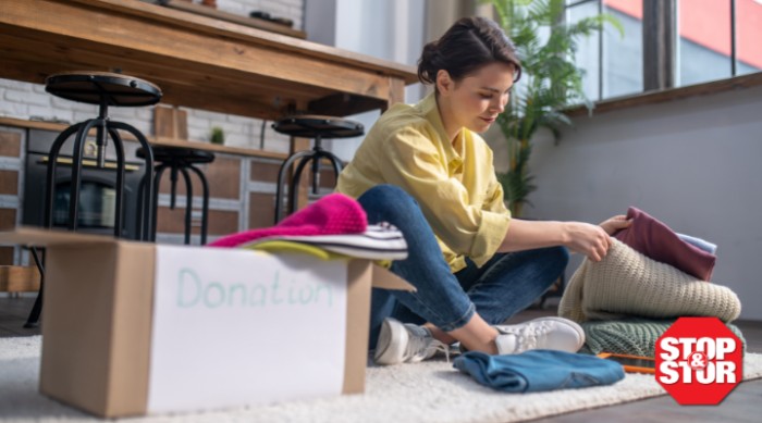 A person sitting on the floor sorting and folding clothes into a donation box in a bright, cozy living space, illustrating the process of decluttering and organizing belongings.