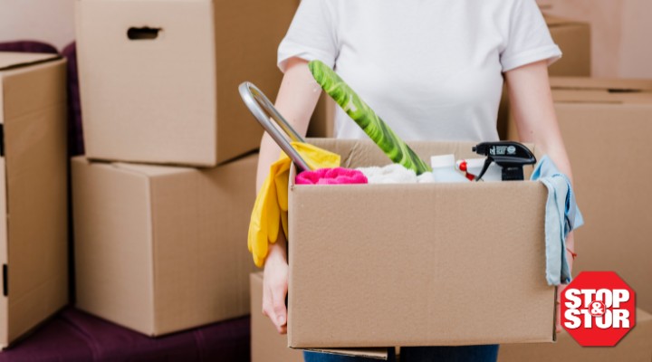 Alt text: Person carrying a box of cleaning supplies and household items while spring cleaning and organizing for self-storage, surrounded by moving boxes in a NYC apartment.